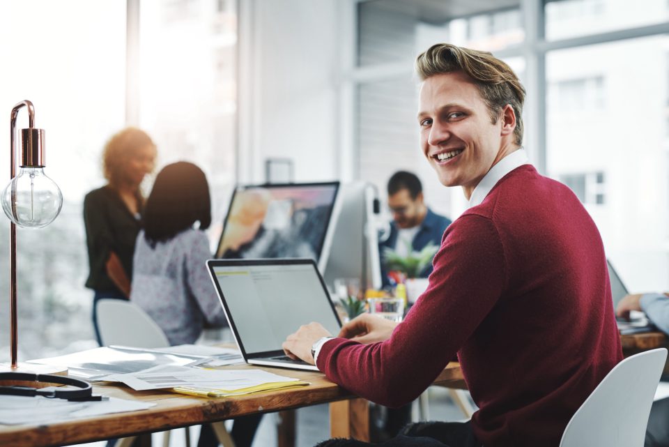 Lachende ondernemer in een rode trui werkt op zijn laptop in een kantoor